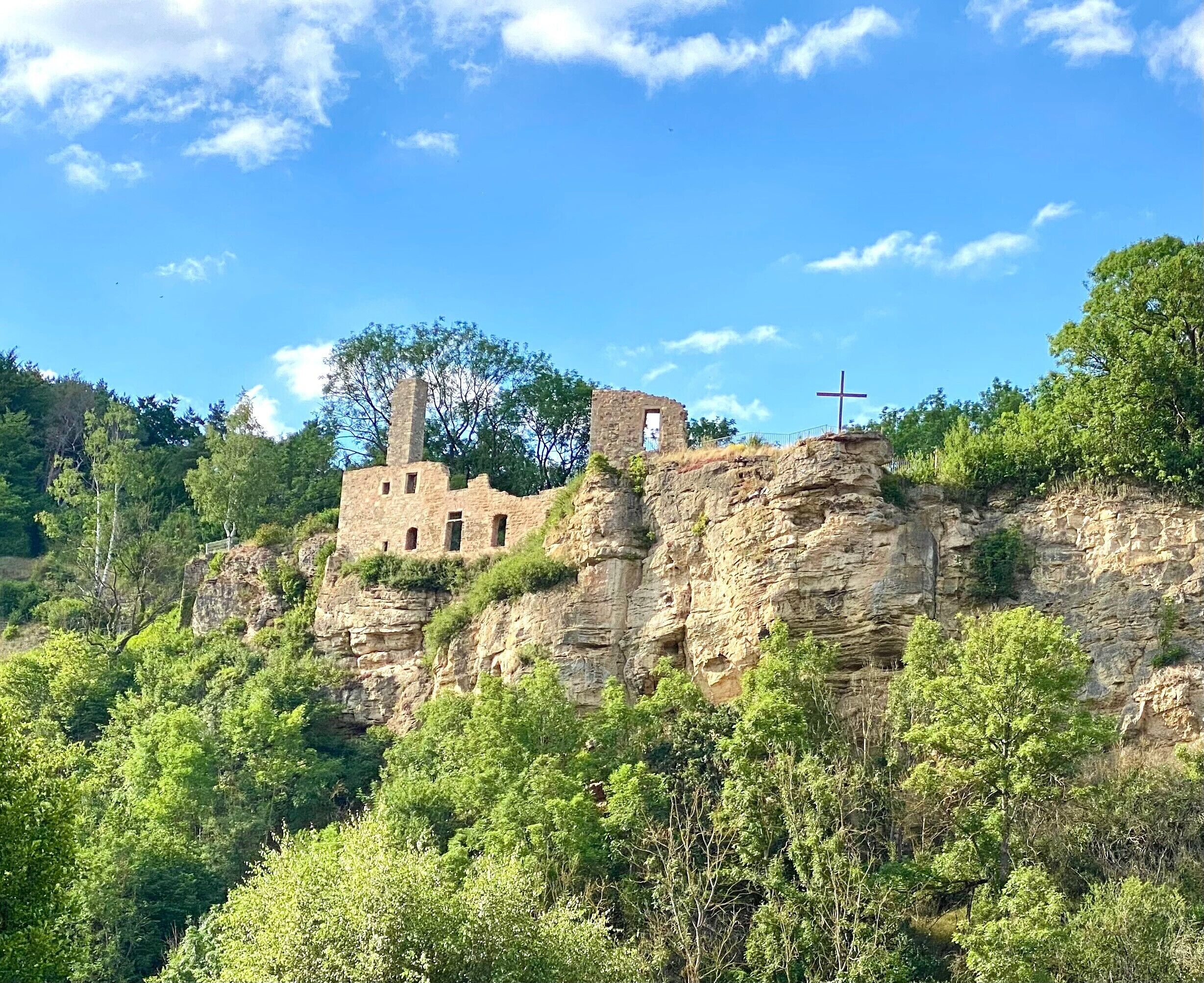 Un bâtiment historique en pierre, situé sur une falaise et surmonté d’une croix, entouré d’une végétation luxuriante.