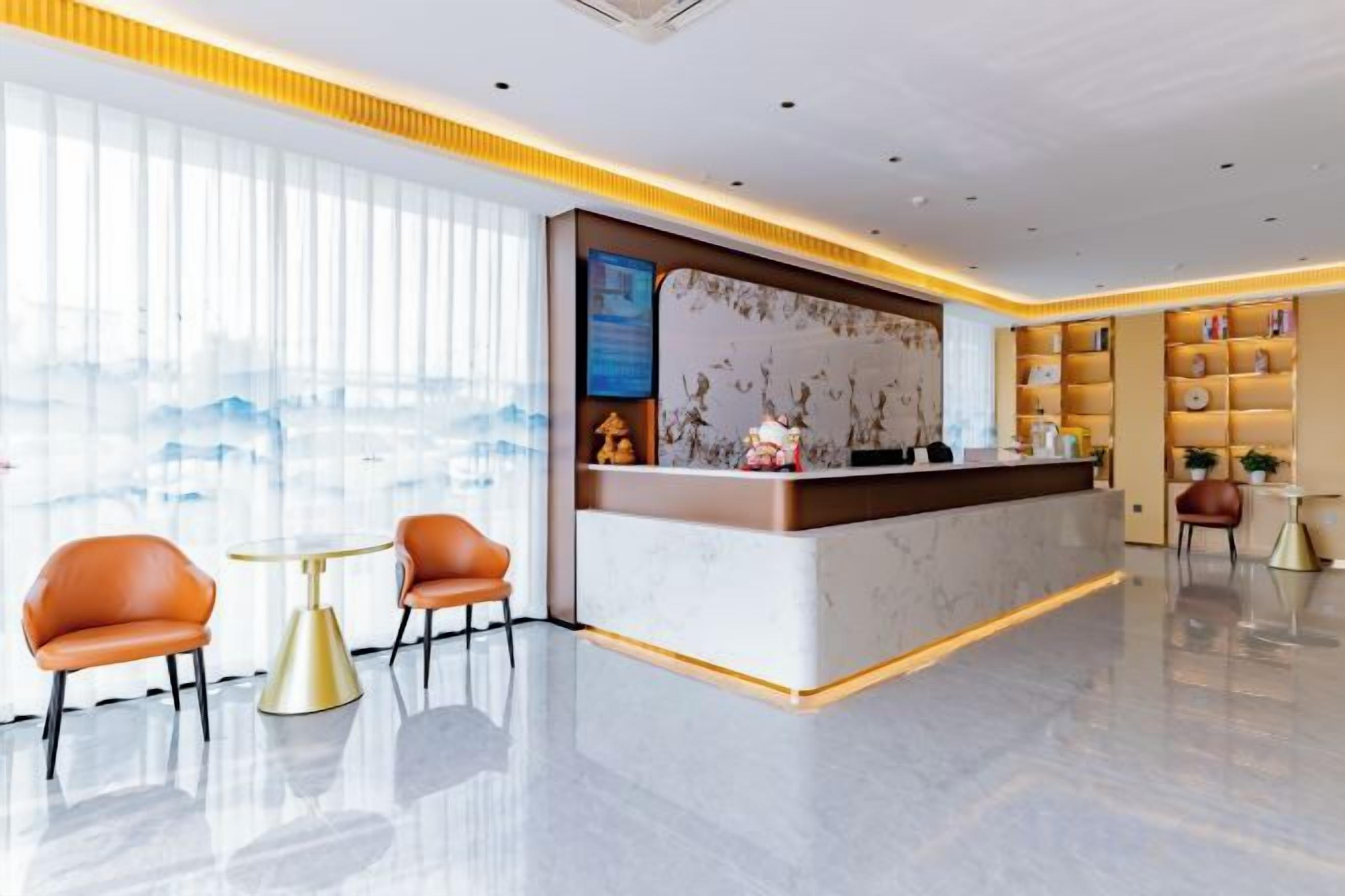 Uma área de recepção de hotel com balcão de mármore, duas cadeiras laranja, uma pequena mesa redonda e uma estante com objetos decorativos.