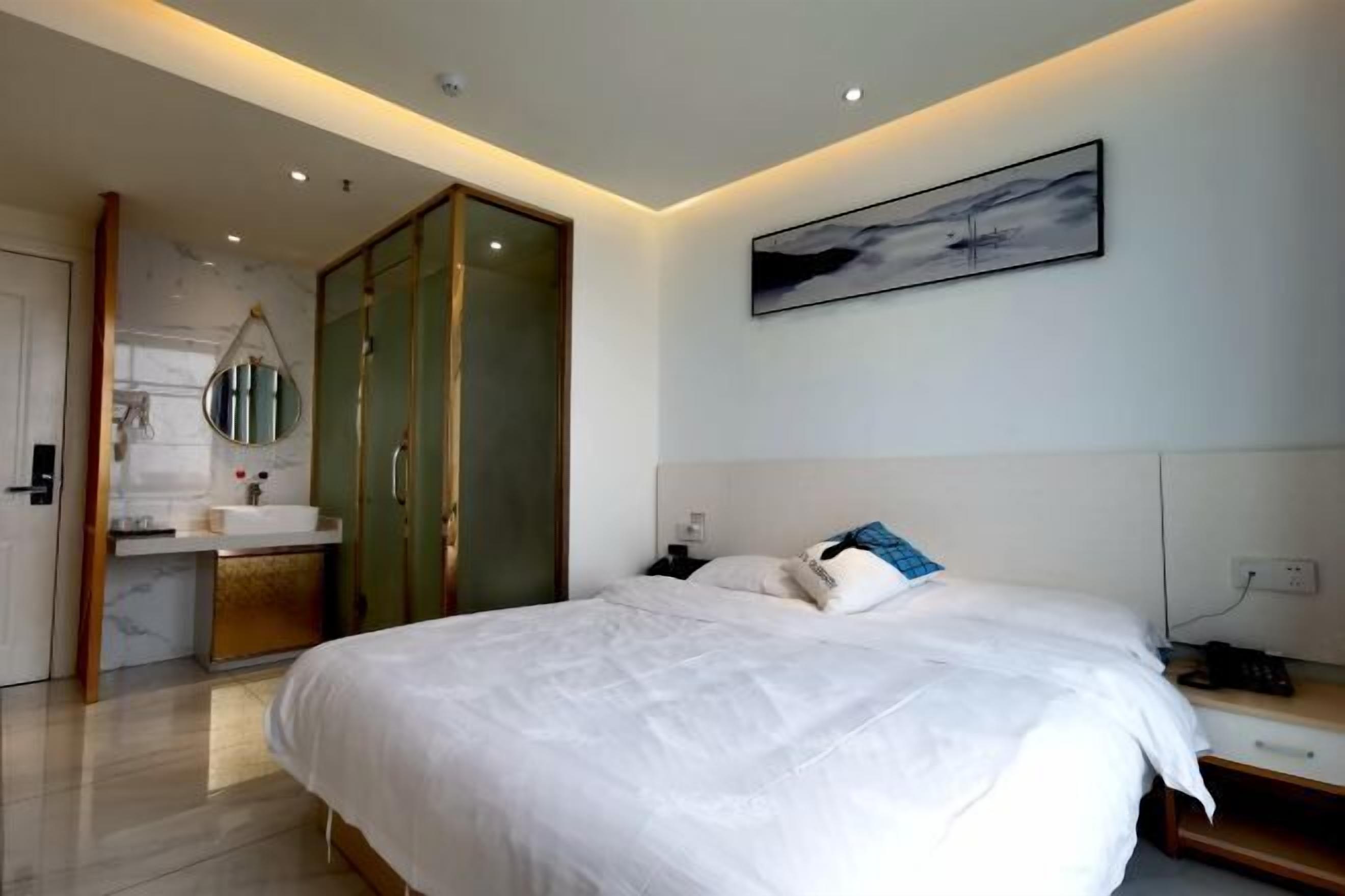 A modern hotel room with a large bed, a bedside table, a nightstand, a wall-mounted artwork, and a bathroom with a sink and mirror.