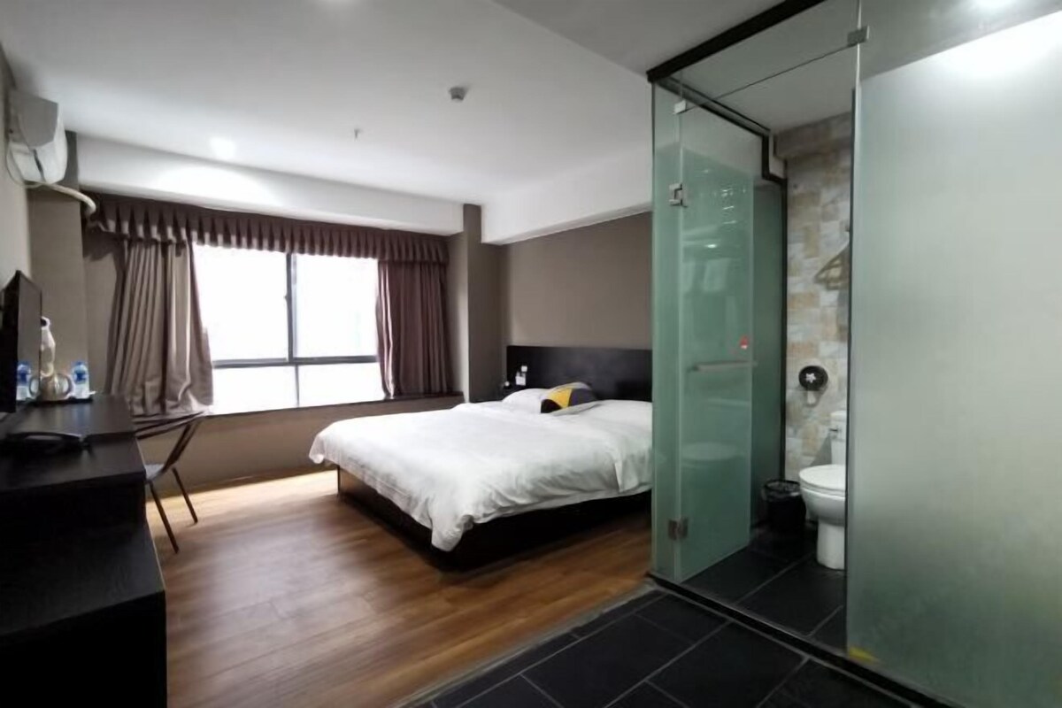 Una habitación de hotel moderna con piso de madera, una cama con ropa blanca, un escritorio con una silla y una zona de ducha con mampara de vidrio.