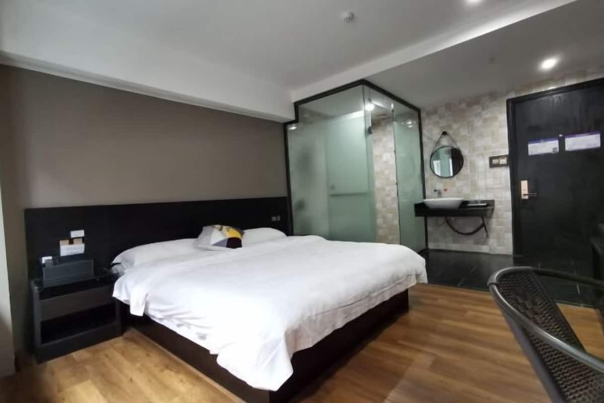 Ein modernes Hotelzimmer mit Holzboden, einem Bett mit weißer Bettwäsche, einer Duschkabine aus Glas, einer schwarzen Tür und einem kleinen Nachttisch mit Lampe.