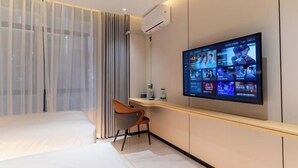 A modern hotel room with a large flat-screen TV mounted on the wall, a bed, a desk with a chair, and a window with sheer curtains. - Huizhou galston hotel (Huizhou)