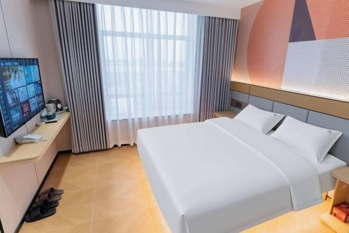 Ein Hotelzimmer mit Bett, Schreibtisch mit Fernseher, Fenster mit Vorhängen und Wand mit geometrischem Muster.
