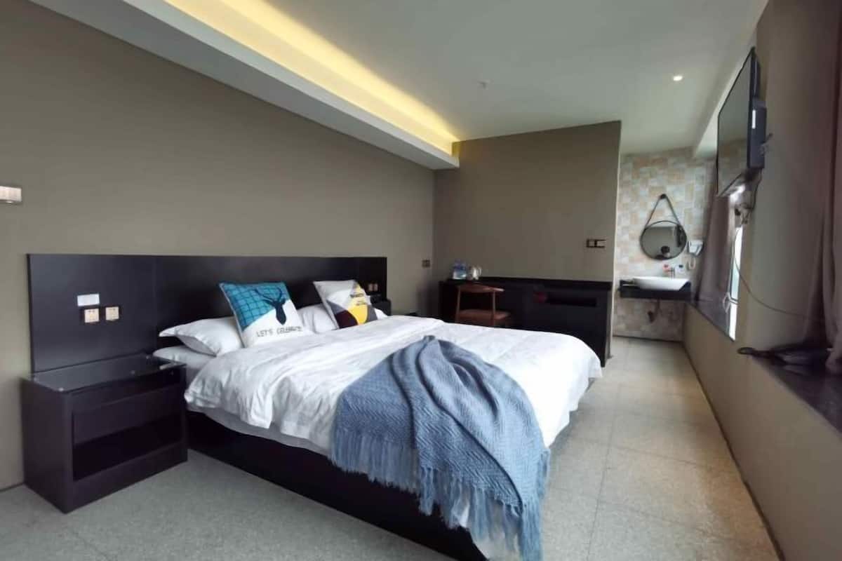 Ein modernes Hotelzimmer mit einem großen Bett, einem Nachttisch, einem fest installierten Fernseher und einem Badezimmer mit Spiegel und Waschbecken.