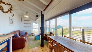A spacious room with a wooden floor, a large window overlooking a scenic view, a wooden dining table, and a cozy seating area with a brown leather sofa. - C24 Rosemary, Riviere Towans (Hayle)