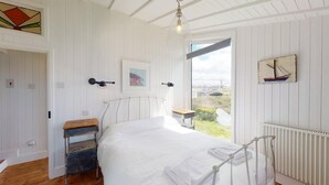 A bedroom with a bed, a small table, a radiator, and a window with a view of the outside. - C24 Rosemary, Riviere Towans (Hayle)