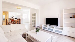 Una sala de estar moderna con un sofá blanco, una mesa de centro con un jarrón de flores, un televisor de pantalla plana sobre un mueble blanco y una estantería.
