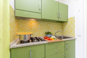 A kitchen with green cabinets, a tiled backsplash, a gas stove, a pot, a cutting board with vegetables, and a sink.