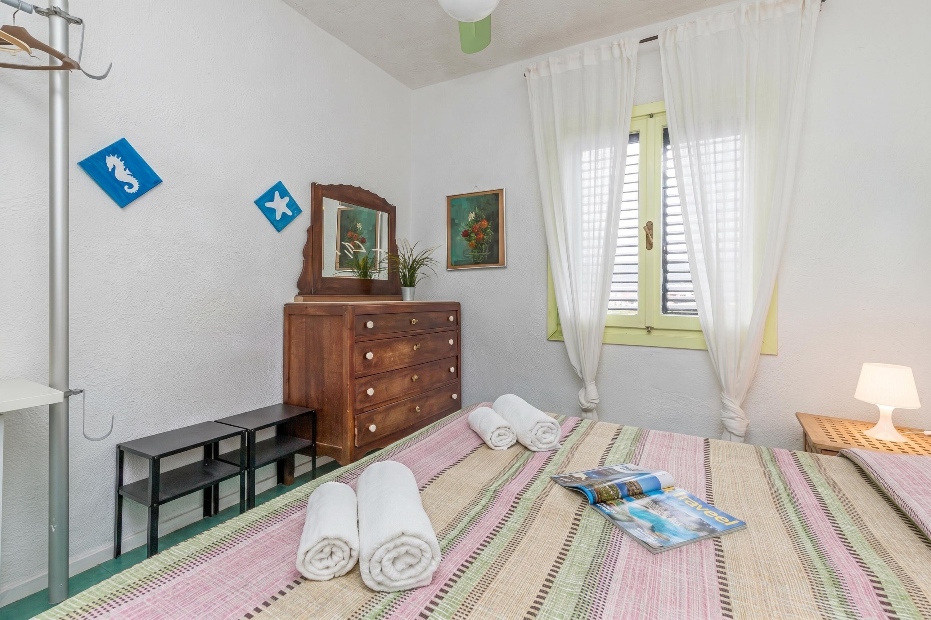 Une chambre à coucher comprenant un lit, une commode, un miroir, une fenêtre avec des rideaux et une petite table avec une lampe.