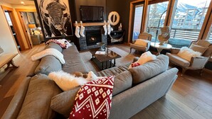 A modern living room with a large gray sectional sofa, a fireplace, and a view of a mountain landscape through large windows. - Iron Mountain Penthouse - World-class Luxury in the Rocky Mountains (Canmore)