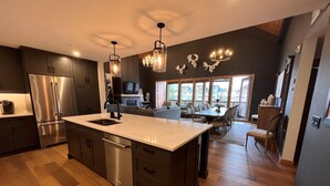 A modern kitchen with dark cabinetry, a stainless steel refrigerator, and a large island with a dishwasher. There is a dining area with a wooden table and chairs, and a chandelier hanging above. - Iron Mountain Penthouse - World-class Luxury in the Rocky Mountains (Canmore)