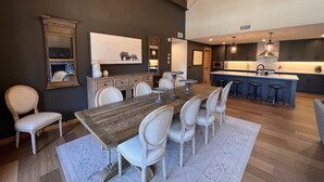 A spacious dining area with a wooden table and white chairs, a kitchen with dark cabinets and a large island, and a chandelier with multiple bulbs. - Iron Mountain Penthouse - World-class Luxury in the Rocky Mountains (Canmore)