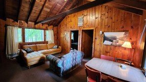 Ein gemütliches Blockhaus-Interieur mit Holzbalkendecke, einer Couch, einem gemusterten Sessel, einem Esstisch mit Stühlen und einem Bild an der Wand.