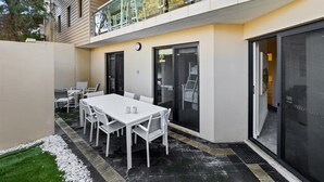 A modern outdoor patio with a white table and chairs, a glass balustrade, and a door leading to an interior space. - Surfside 1—An Unforgettable Yallingup Experience (Yallingup)