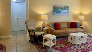 Uma sala de estar com um sofá bege, uma cadeira de balanço, um banquinho branco, um tapete colorido e um baú de vime.