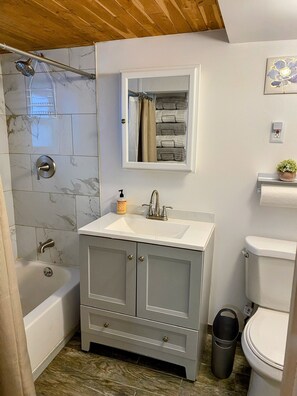 A bathroom with a bathtub, a sink with a mirror above it, and a toilet. There is a shower curtain rod above the bathtub, and a trash can is visible near the toilet. - King Bed & In-Unit Laundry! (Chicago)