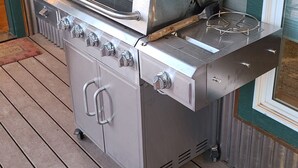 En udendørs grill i rustfrit stål med fire brændere, ovn og sidebrænder, placeret på en træterrasse.