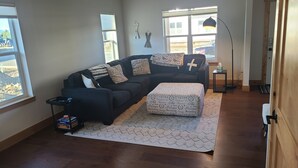 En stue med en mørk sofa, en polstret skammel med mønster og et lille sofabord. To vinduer giver naturligt lys, og der er en lampe på sofabordet.