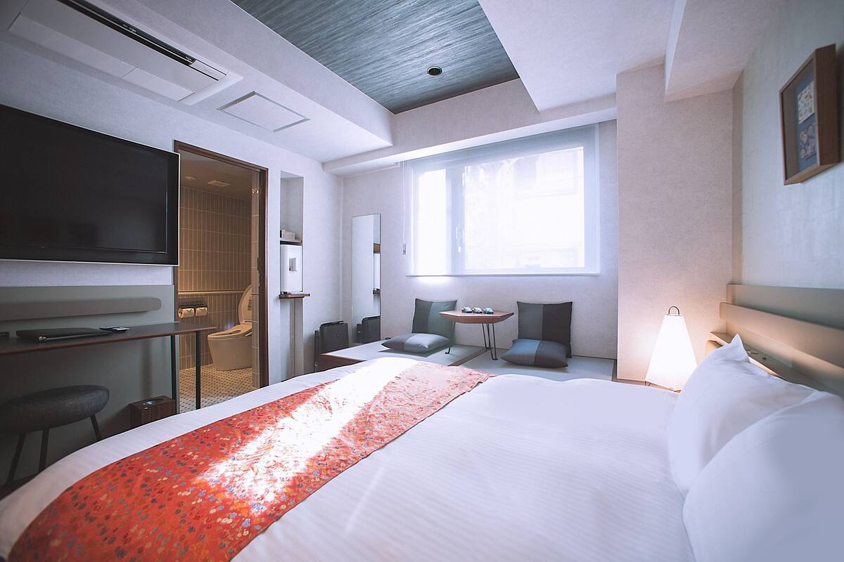 Quarto de hotel com uma cama grande, uma secretária com televisão de tela plana, uma pequena mesa com cadeiras e uma janela que permite entrada de luz natural.