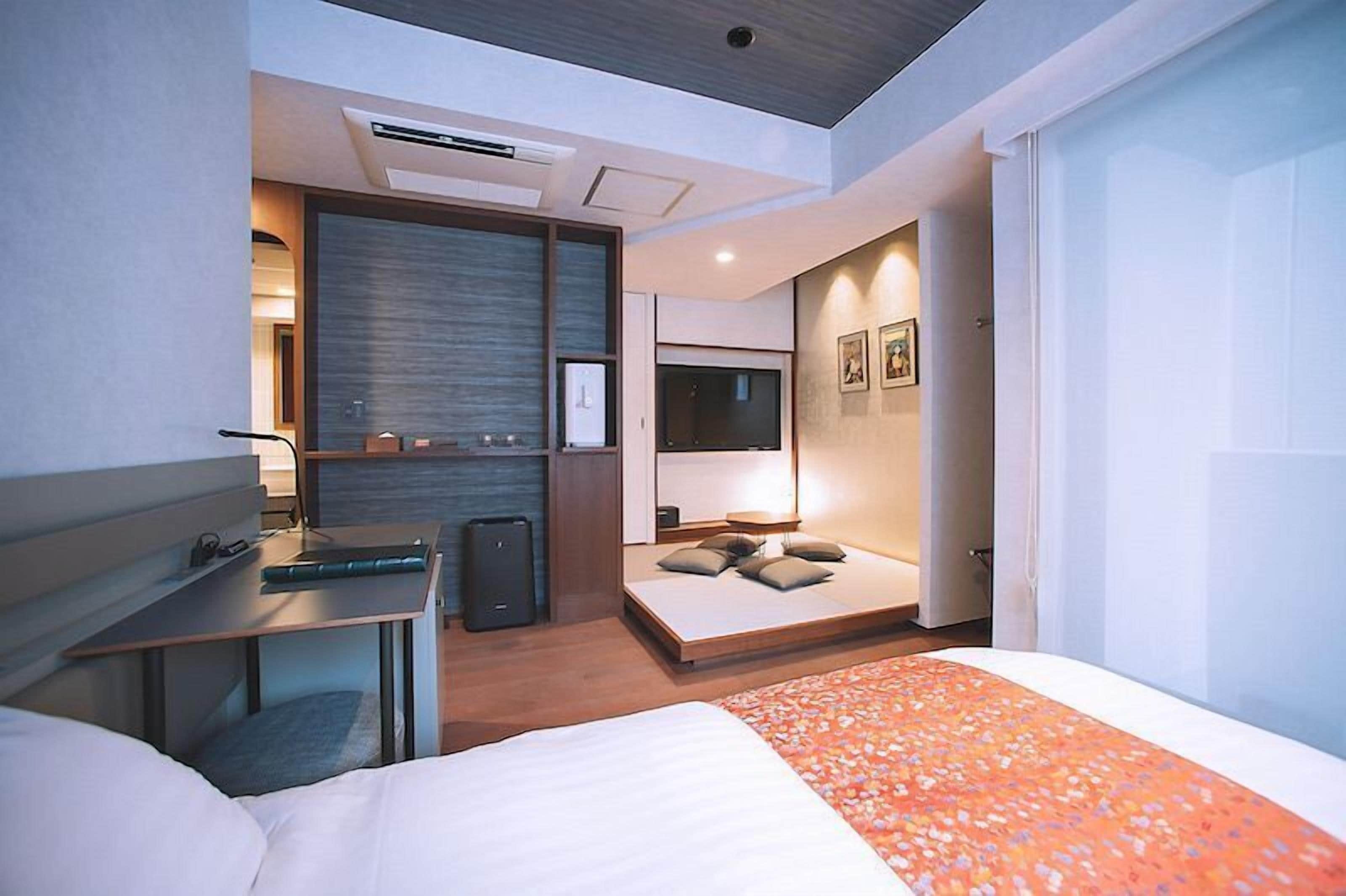 A modern hotel room with a bed, a desk, a seating area, and a TV.