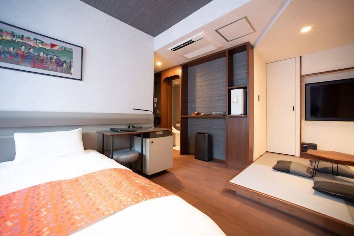 Quarto de hotel com cama, escrivaninha, cadeira, televisão e uma pequena copa.