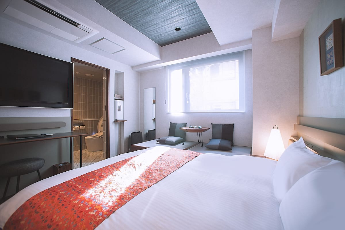 Quarto de hotel com uma cama grande, uma secretária com televisão de tela plana, uma pequena mesa com cadeiras e uma janela que permite entrada de luz natural.