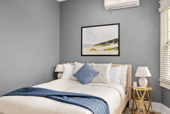 Une chambre à coucher avec un lit, des oreillers, une table de chevet avec une lampe, un tableau encadré au mur et une fenêtre avec des stores.