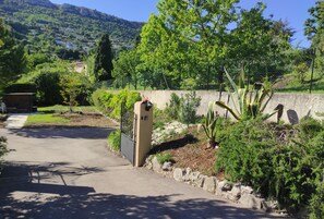 Une entrée fermée par un portail et bordée d’un mur de pierre, une végétation luxuriante et une vue dégagée sur un paysage montagneux en arrière-plan.