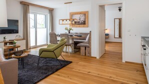 Een moderne woonkamer met houten vloer, een groene fauteuil, een eethoek met tafel en stoelen, een aan de muur bevestigde televisie en een keuken met aanrecht en kastjes.