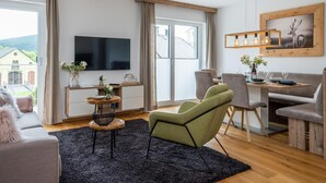 Een moderne woonkamer met een flatscreen televisie, een eethoek met een houten tafel en stoelen, en een groot raam met uitzicht op een gebouw en groen.