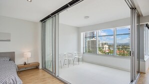 A modern bedroom with a large sliding glass door leading to a balcony, a bed with a grey and white polka dot duvet, a bedside table with a lamp, and a view of city buildings through the window. - Sky Point Tower - Private Apartments (Surfers Paradise)