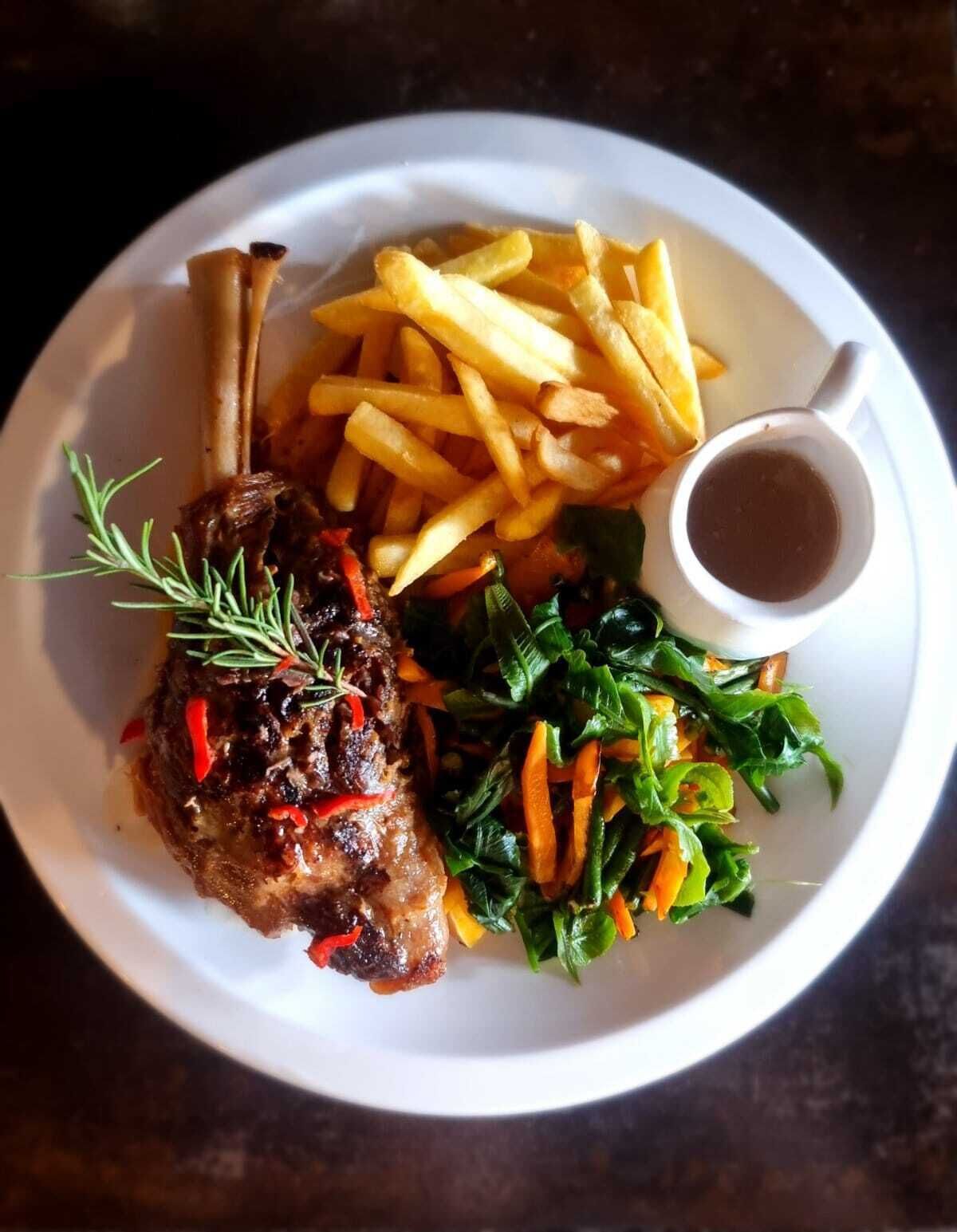 Une assiette comprenant un jarret d’agneau, des frites, un assortiment de légumes verts et une petite portion de sauce.