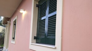 Ein Gebäude mit rosafarbenem Anstrich, einem Fenster mit grünen Rollläden und einem Blumentopf daneben.