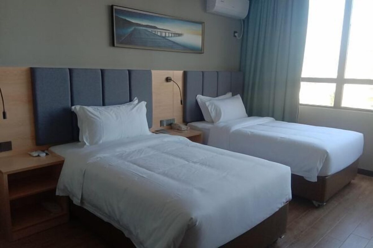 A hotel room with two single beds, a wooden nightstand, a desk, a lamp, and a painting on the wall.