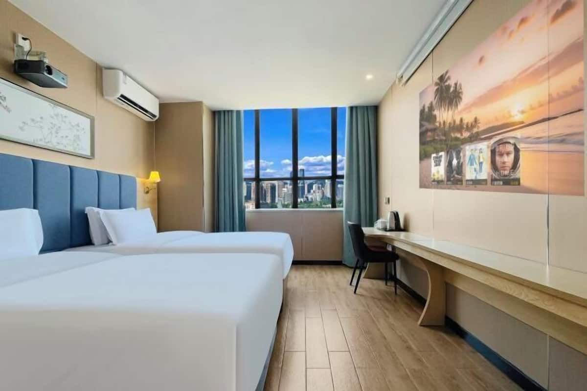 Habitación de hotel con dos camas, un escritorio, una silla, un ventanal con vistas a la ciudad y un equipo de aire acondicionado.