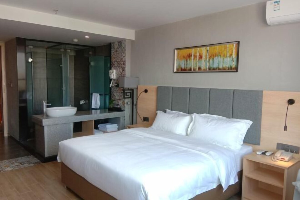 A modern hotel room with a large bed, a bathroom with a glass shower enclosure, a sink, and a bedside table with a lamp and phone.