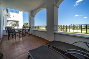 Ein überdachter Außenbereich mit Tisch und Stühlen, Blick auf einen Golfplatz und ein weißes Gebäude im Hintergrund.