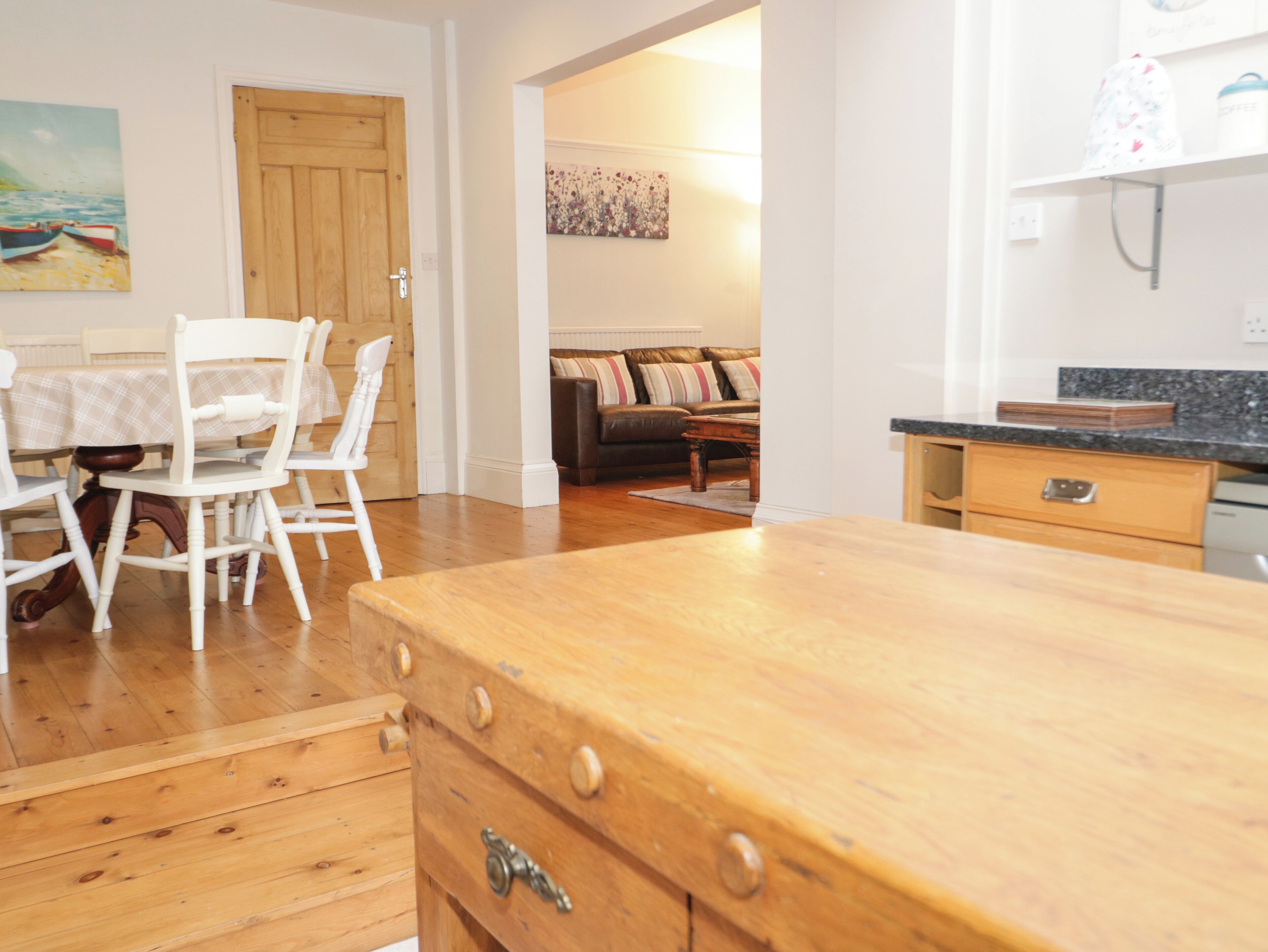A kitchen with wooden cabinets, a dining area with white chairs, and a sofa in the background.