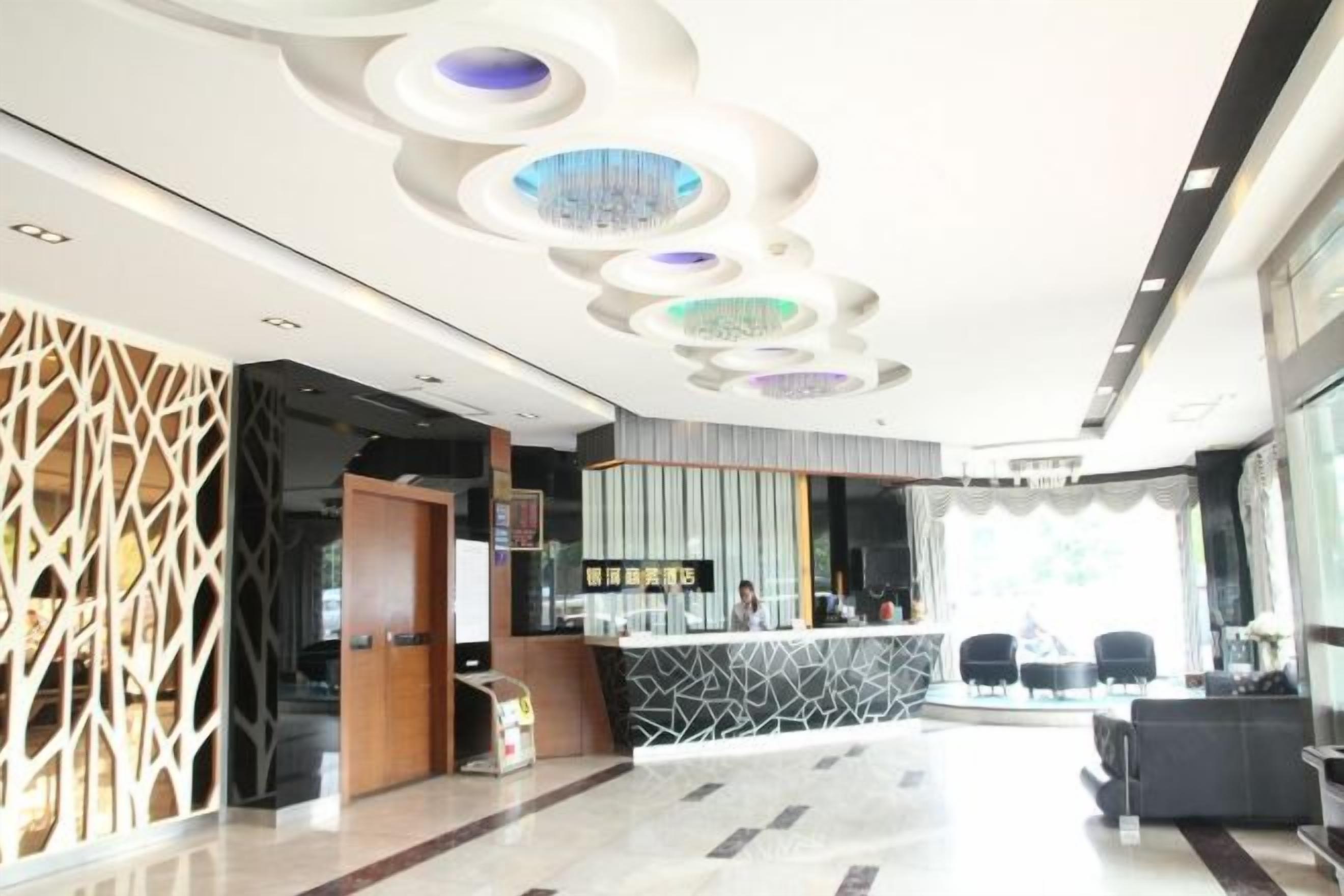 A modern hotel lobby with a reception desk, seating area, and decorative ceiling lights.