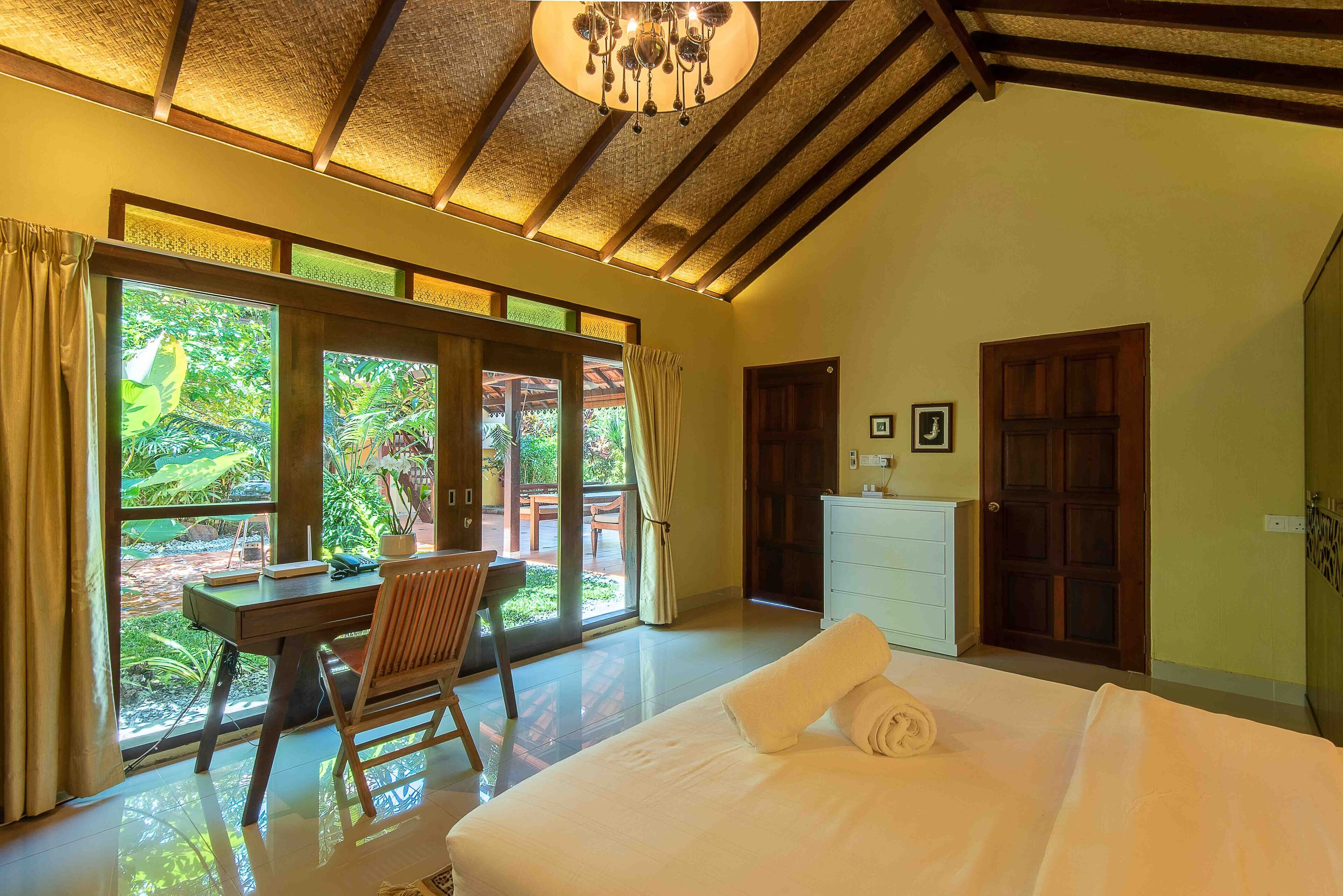 A bedroom with a bed, a desk, and a chair. There are large windows and a door.