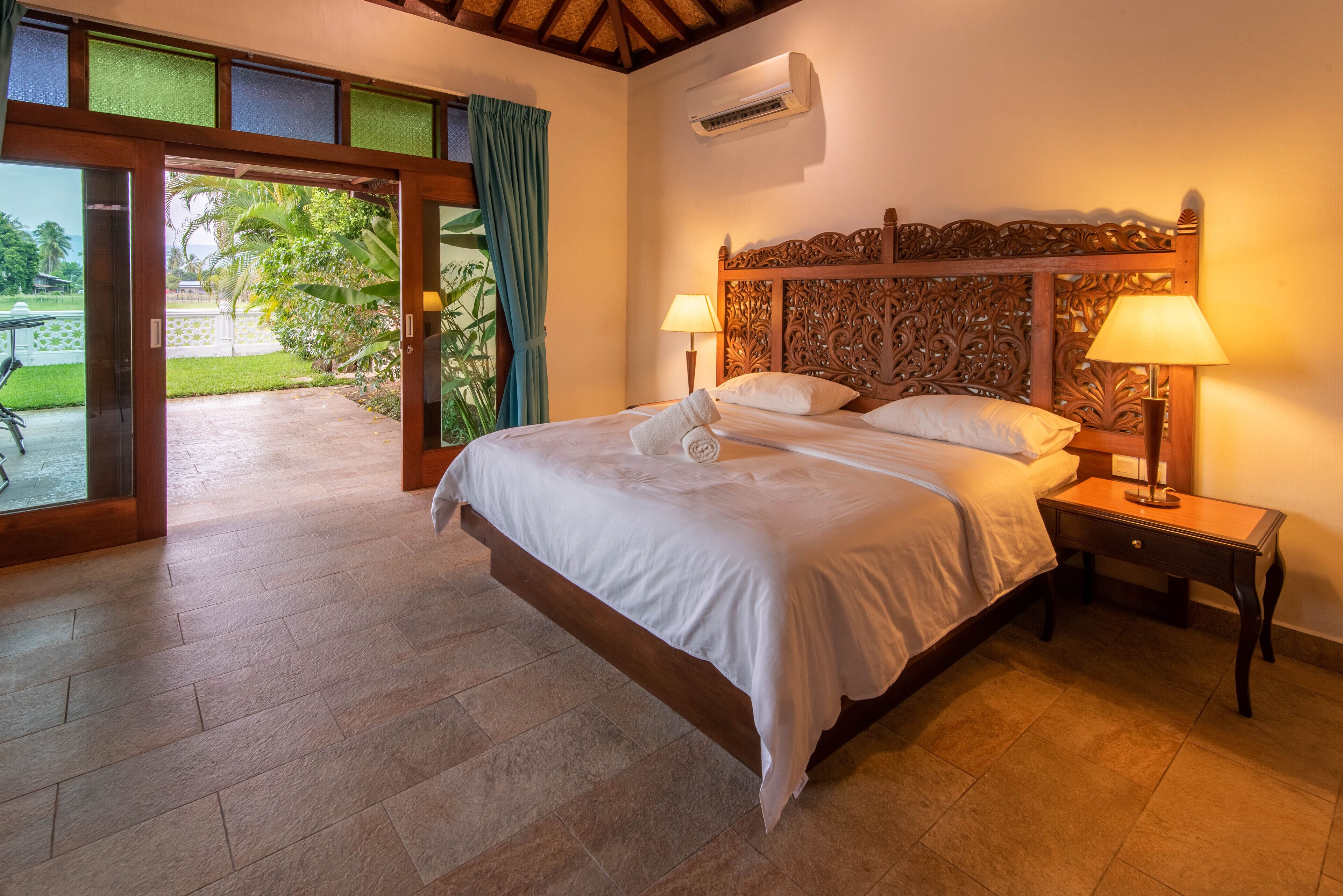 Ein Hotelzimmer mit einem großen Bett, zwei Nachttischlampen, einem Holz-Kopfteil und Blick auf einen Garten durch eine Glasschiebetür.