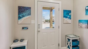 A hallway with a white door, blue artwork on the walls, a small white table with a blue bin, and a glass display case with blue bins. - 1486 - Blue Retreat - Whalehead Corolla Escape w/ Pool & Hot Tub (Corolla)