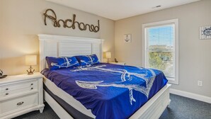 A bedroom with a bed, two bedside tables, a window with blinds, and wall decorations. - 1486 - Blue Retreat - Whalehead Corolla Escape w/ Pool & Hot Tub (Corolla)