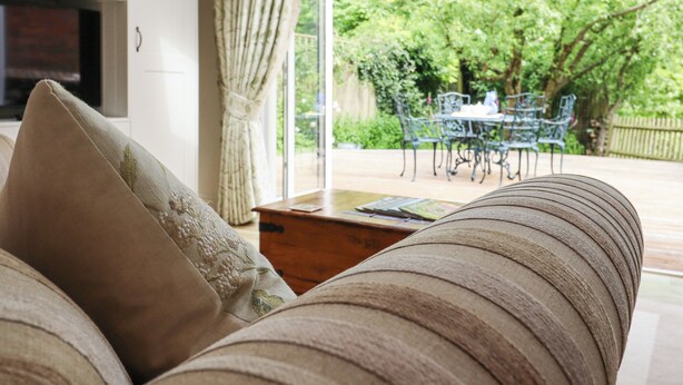 A cozy living room with a sofa, a wooden coffee table, and a bookshelf. A large window with curtains reveals a garden with a table and chairs outside. - Oast Cottage (Maidstone)