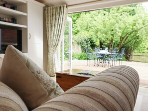 Uma sala de estar aconchegante com sofá, mesa de centro de madeira e estante. Uma janela grande com cortinas mostra um jardim com mesa e cadeiras no exterior.