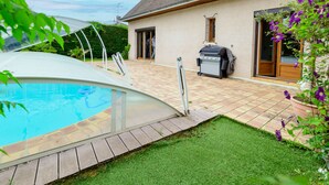 Une piscine entourée de parois en verre, un barbecue et une maison dotée d’un patio carrelé.