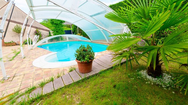 Une piscine située sous une structure en verre de type serre, entourée d’une terrasse en briques et d’une végétation luxuriante.