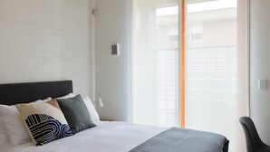 Ein Hotelzimmer mit Bett, Kissen, Nachttisch, Lampe und Blick ins Freie durch ein Fenster.