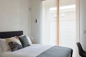 A hotel room with a bed, pillows, a bedside table, a lamp, and a view of the outside through a window. - Just My Type Richmond Retreat with Parking (Richmond)