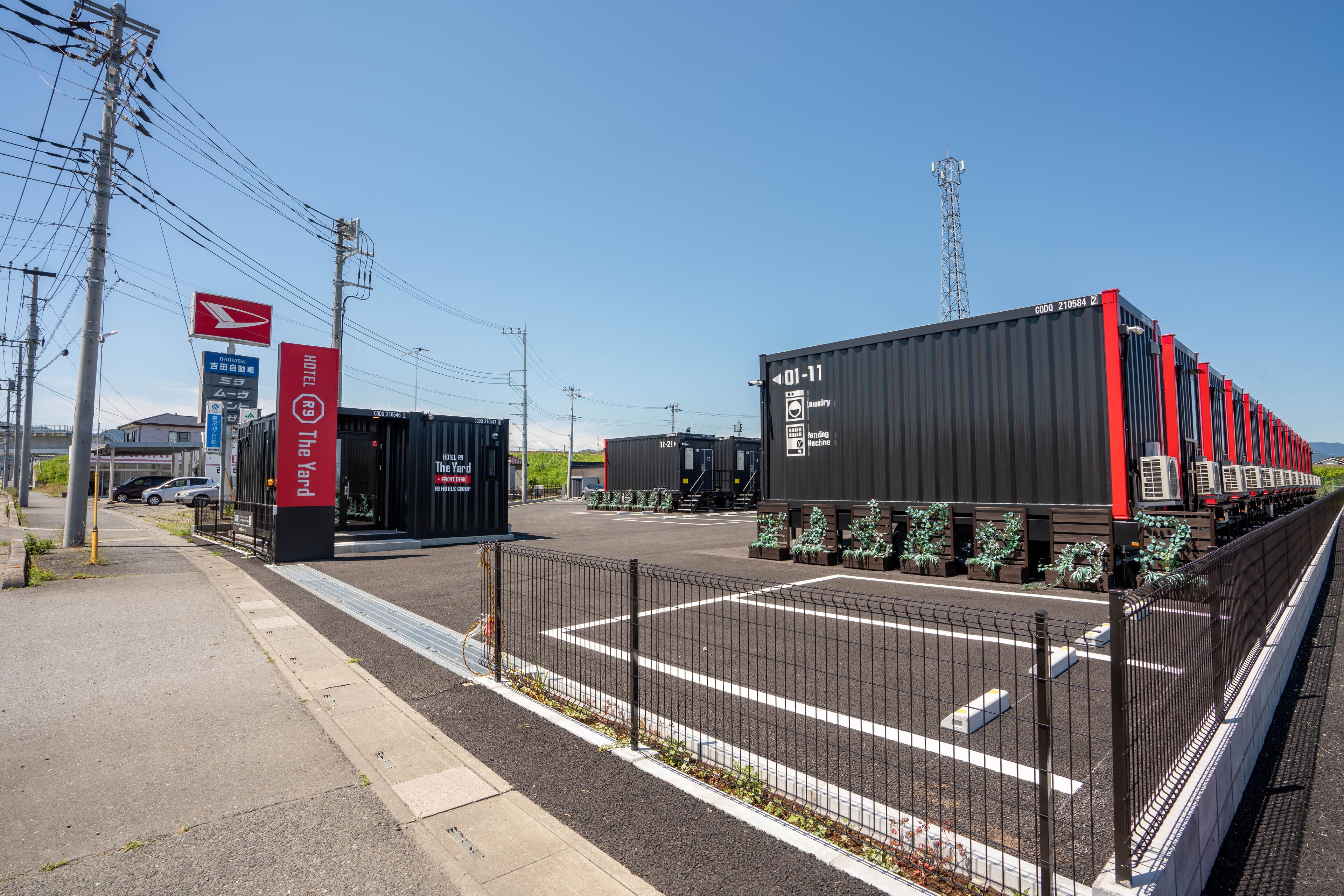 A hotel entrance with a red sign, a parking lot, and shipping containers in the background.